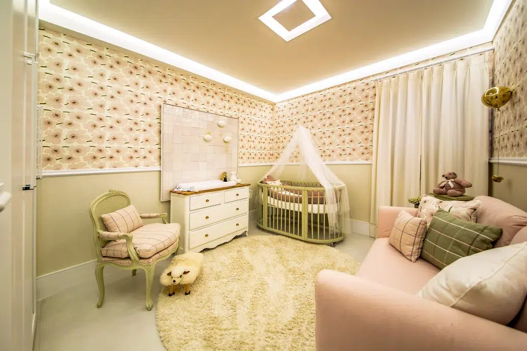 Quarto de uma bebé com um  sofá de cor rosa, uma cuna em tons rosas e  brancos, uma meia parede horizontal lisa e a outra com desenhos.