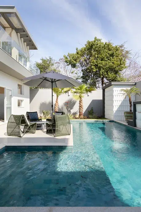 piscina em um jardim de cores neutros, sobrado de paredes brancas, cadeiras e guarda-sol cinza. 