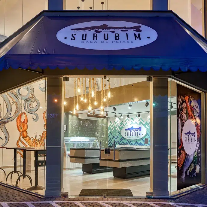 Fachada de um mercado de peixes com paredes de vidro, um toldo azul com o logo da loja, na lateral um letreiro iluminado com logo da marca.