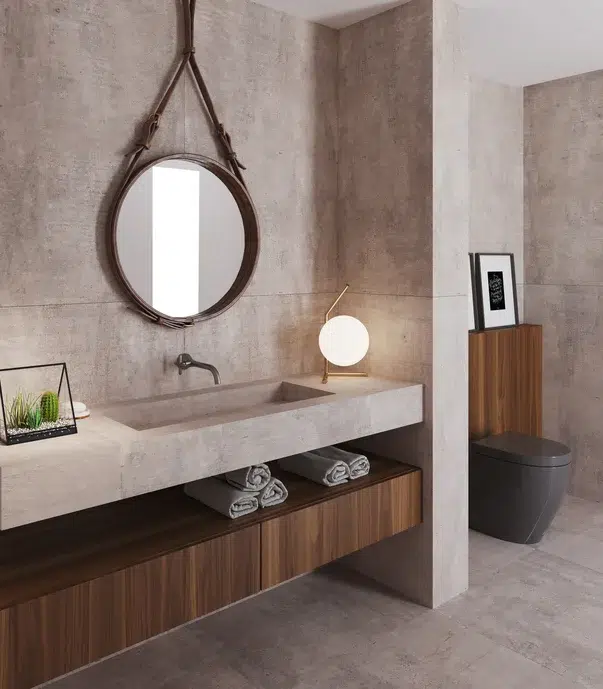 Banheiro cinza externo com revestimento de porcelanato, um espelho redondo acima da pia, uma bola de luz, e dois cactus decoração.