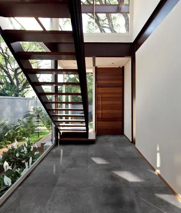 Área de entrada de uma casa com a porta de madeira e as paredes de vidro, com uma escada de madeira larga e comprida.