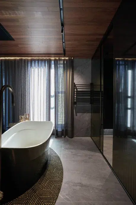 Banheiro cinza de madeira com uma banheira no meio e umas cortinas na janela deixando entrar pouca luz.