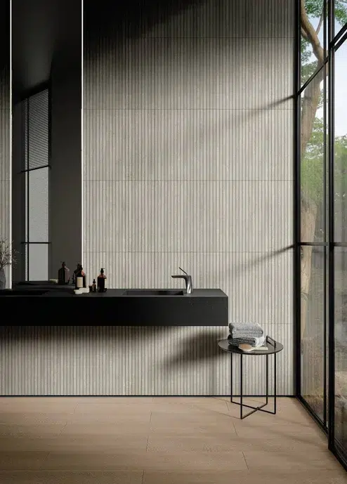 Banheiro cinza com revestimento de madeira, pia em cor preta, um banco pequeno e baixo com toalhas e um espelho.