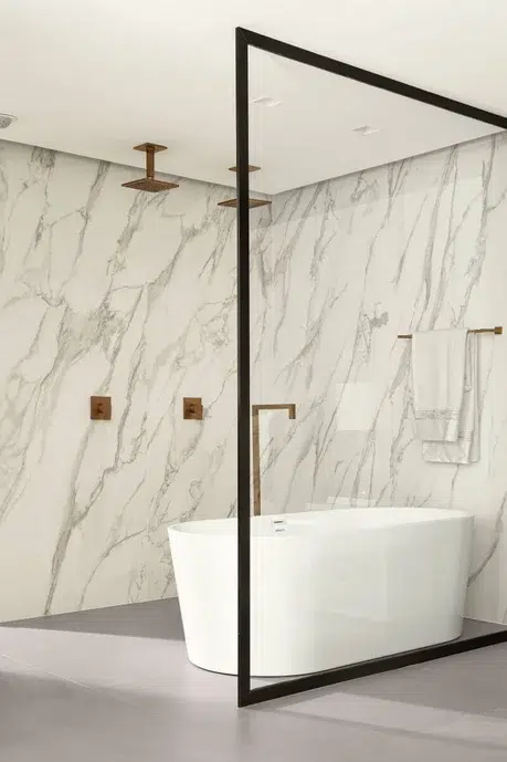 banheiro revestido com mármore branco, uma banheira branca e uma parede de vidro com bordas pretas.