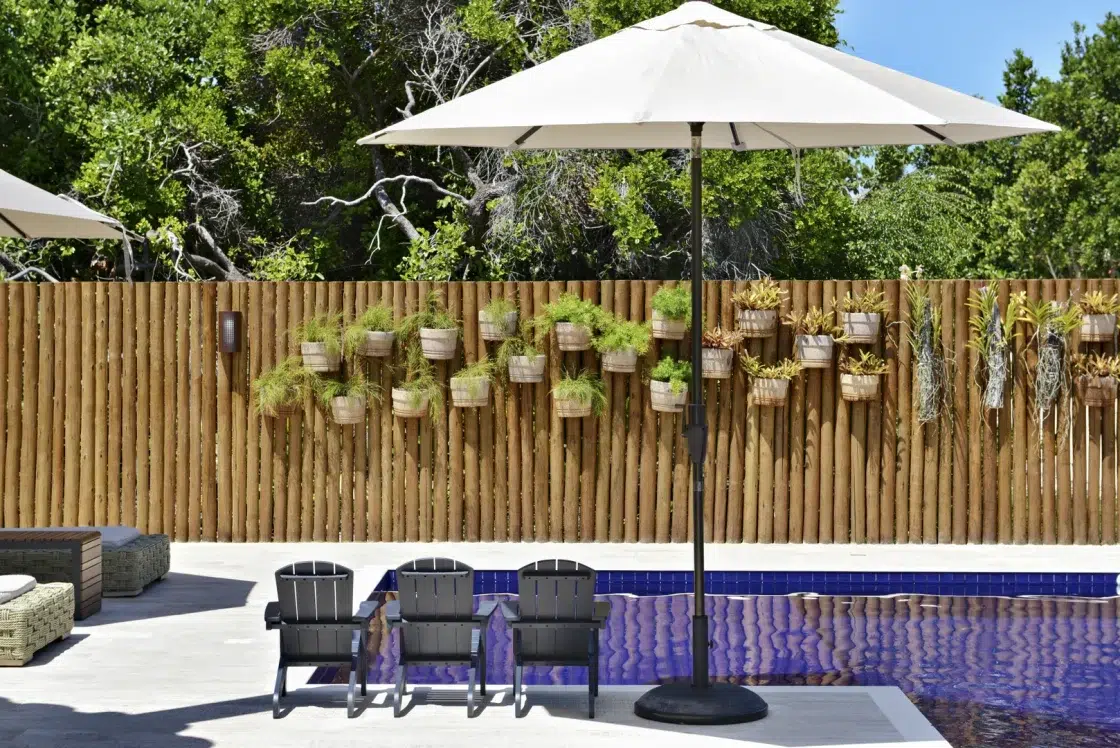 Piscina em L com três cadeiras de madeira ao lado e um guarda-sol. Ao fundo, uma cerca de madeira com vasos de plantas suspensos.