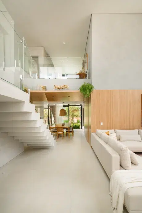 Sala com ambiente integrado com escada revestida em porcelanato, sofás brancos, parede que imita madeira e mesa de jantar de madeira.