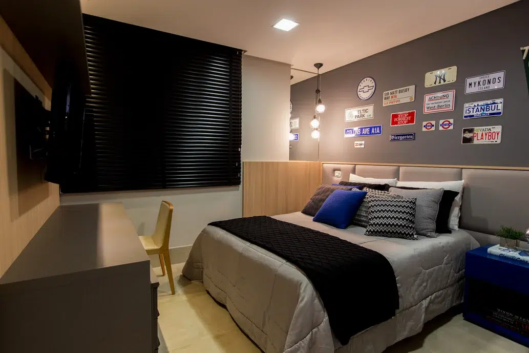 Quarto masculino com decoração de placas coladas na parede e uma cama grande cheia de almofadas em cores neutros.