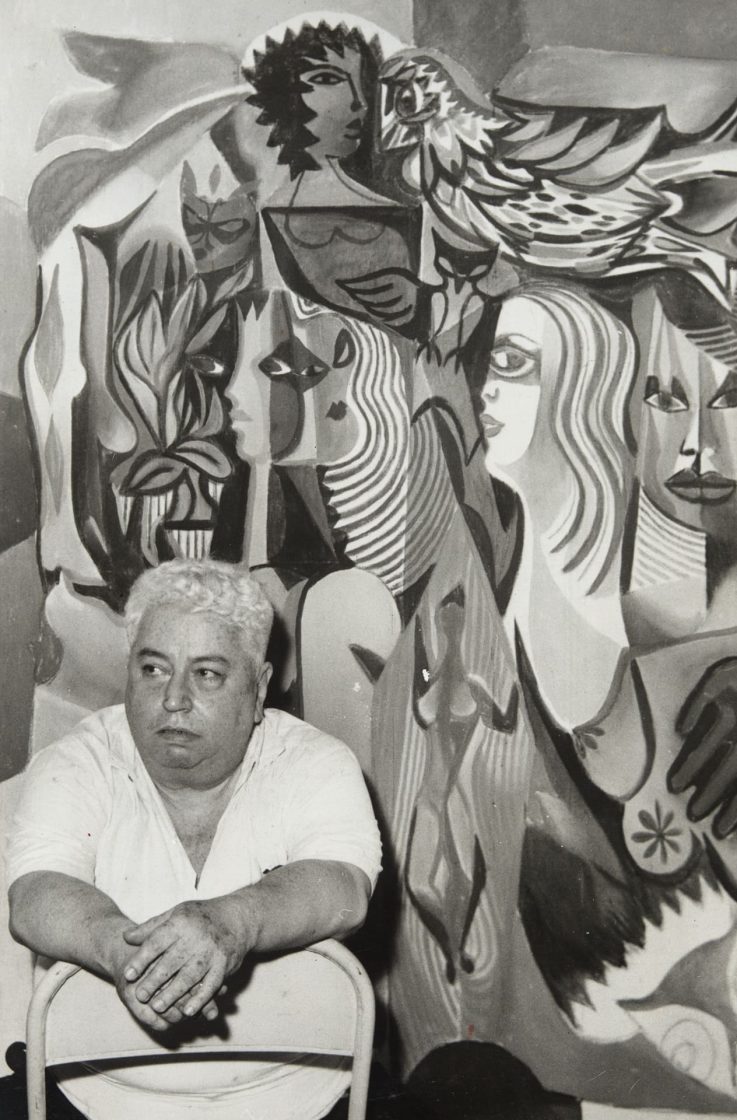 Fotografia de Di Cavalcanti sentado diante de um mural com figuras estilizadas e elementos da estética modernista brasileira
