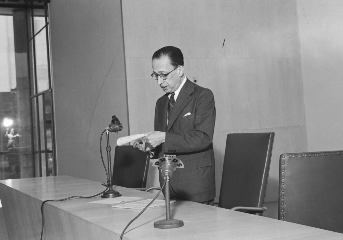 Fotografia de Manuel Bandeira lendo um texto diante de microfone em mesa de conferência durante evento cultural no século 20