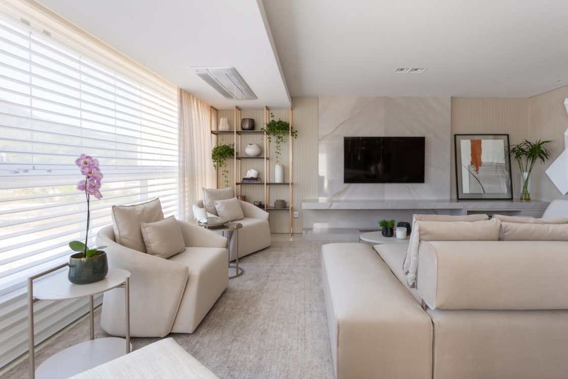 Sala de estar com revestimentos de cor clara, sofá fendi, mesas laterais e estante com metal dourado