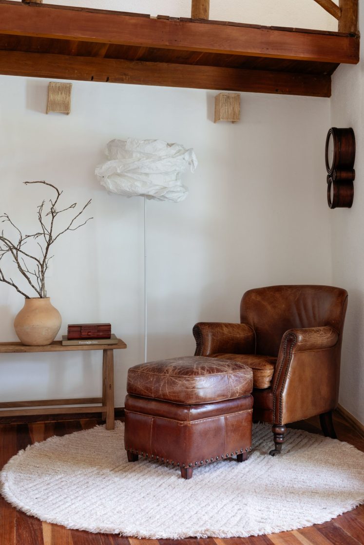Sofá de couro caramelo e luminária branca ao lado, mesa lateral de madeira e vaso de cerâmica 