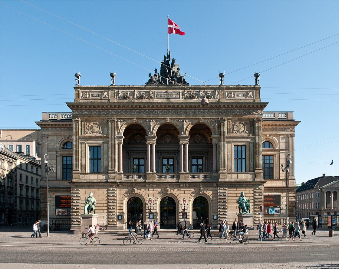 Real Teatro Dinamarquês, edifício neoclássico, bandeira da Dinamarca hasteada e pessoas caminhando