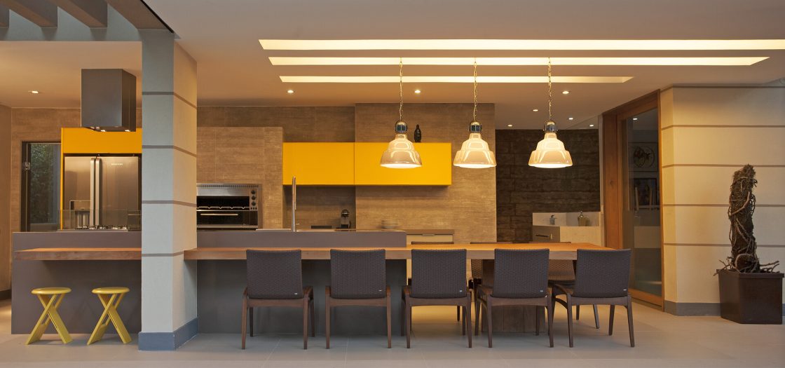 Cozinha gourmet com pontos de cor nos armários amarelos, pendentes e bancada integrada com área de refeição