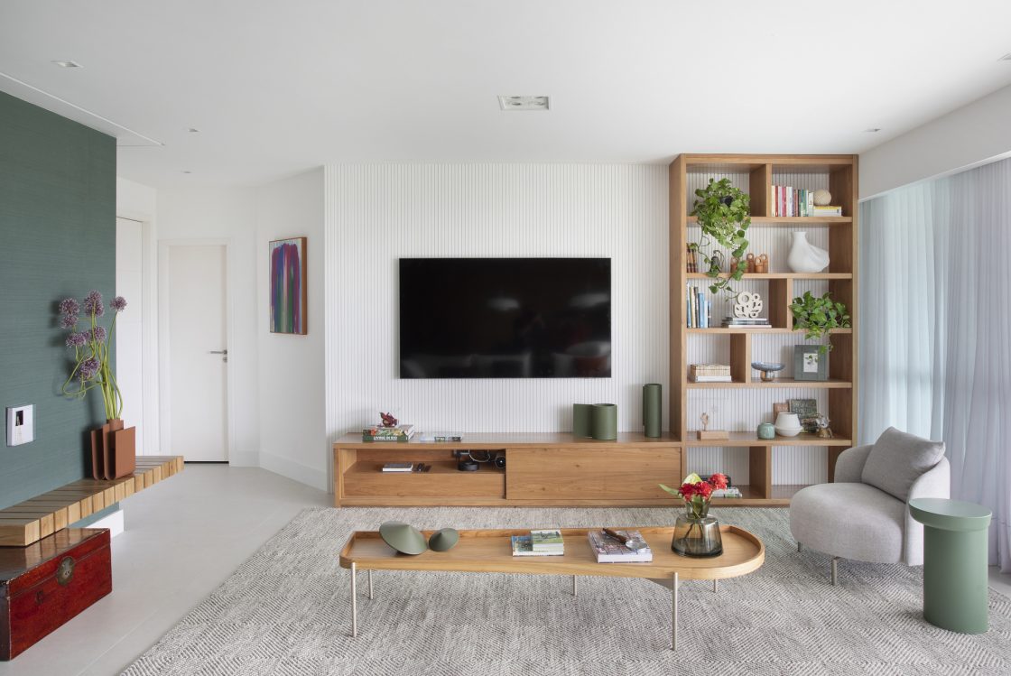 Sala de TV em tons claros com móveis em madeira e planta jiboia no alto da estante