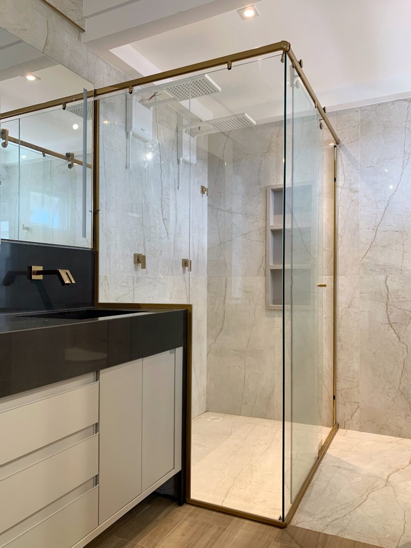 Banheiro com gabinete em preda escura, box com mármore de tons claros e detalhes em dourado