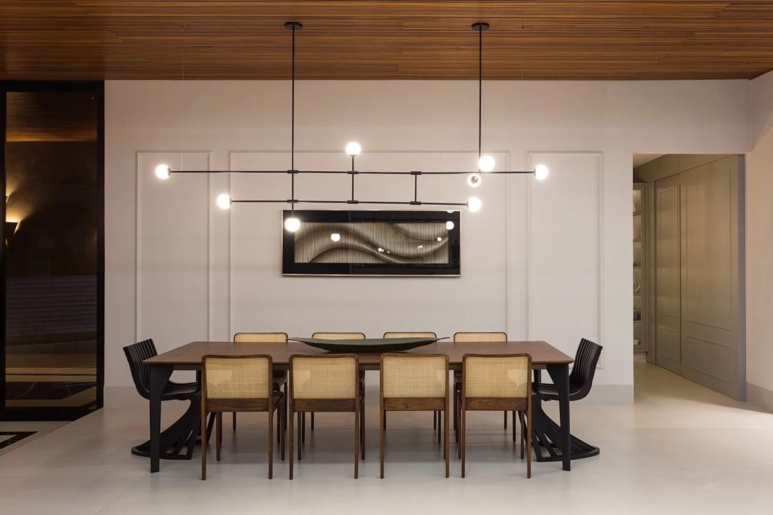 Sala de jantar com paredes claras, boiserie e um quadro encaixado na moldura. Ao centro, há uma mesa em madeira com cadeiras 