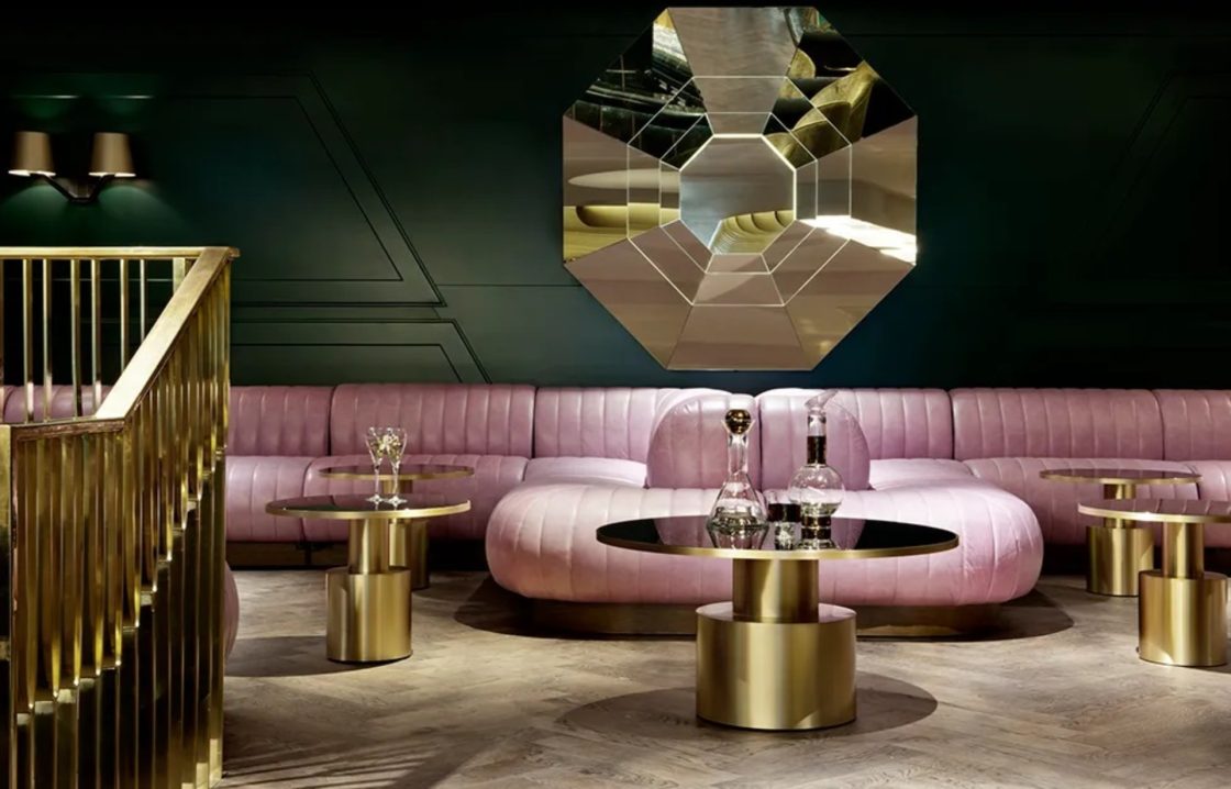 Ambiente no Mondrian Hotel, com parede em tom esverdeado, sofá em rosa, espelho geométrico na parede e detalhes em dourado