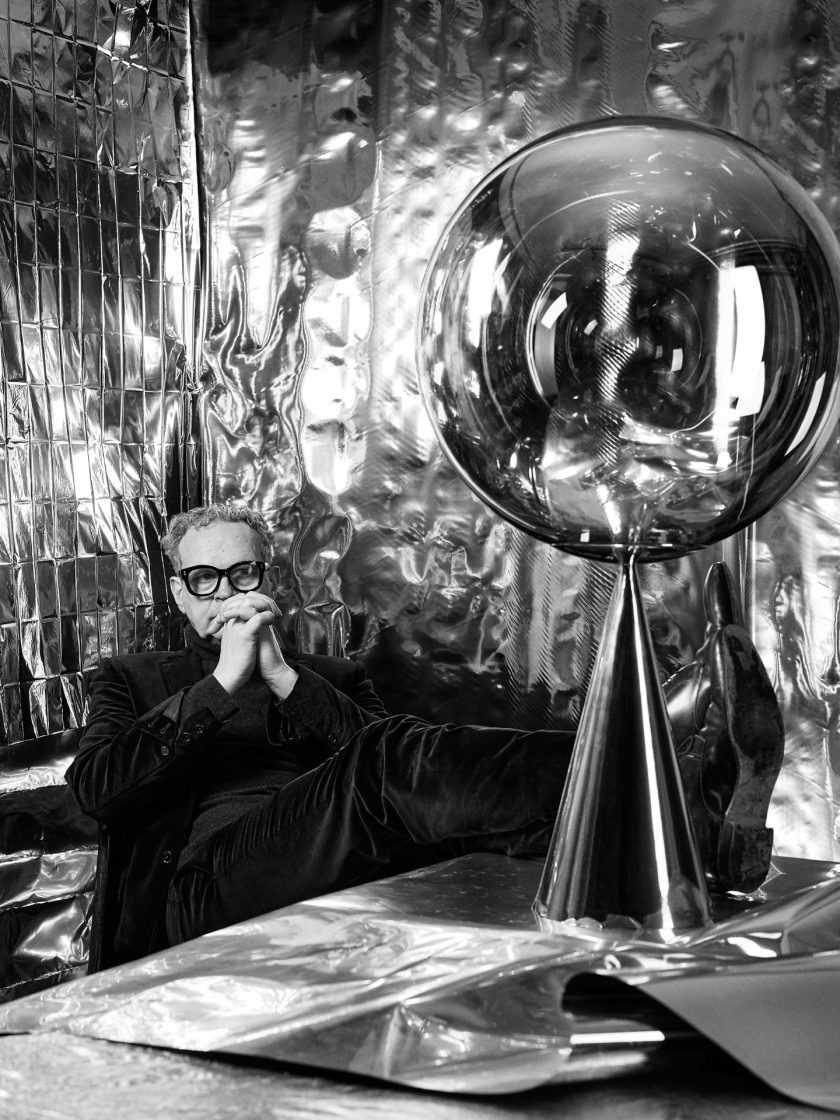 Retrato em preto e branco do designer Tom Dixon, que aparece sentado, com roupas pretas e óculos. Ele está com as mãos cruzadas. 