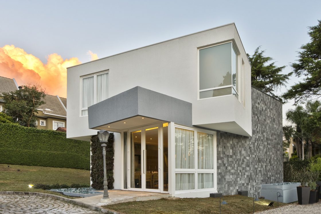 Casa em tons de branco e cinza que traz segundo pavimento com bicos modernos, janelas e portas de vidro