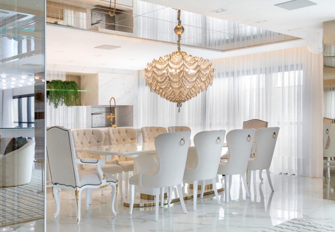 Sala de jantar com parede e móveis brancos, grande lustre ao centro da mesa, que inclui 10 cadeiras com estilo provençal