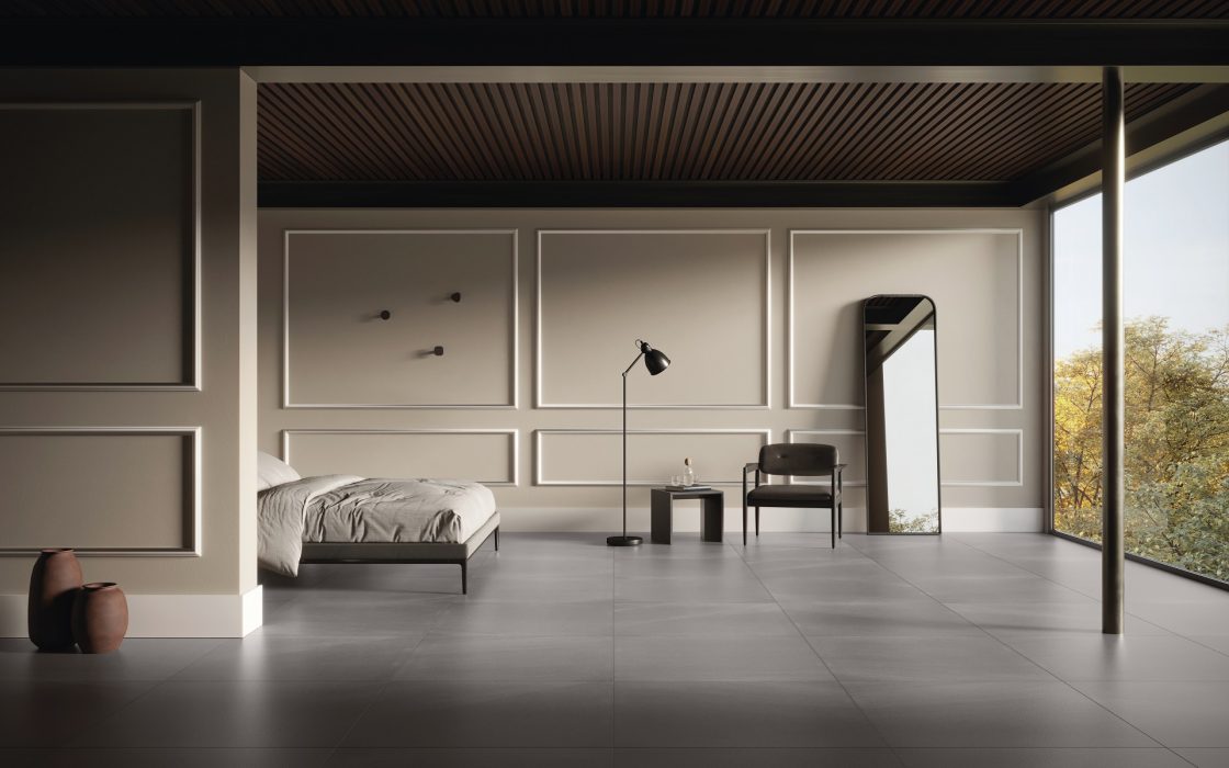 Quarto com parede cinza, com molduras estilo boiserie, cama à esquerda, um espelho, uma luminária de piso, uma poltrona e mesa de apoio