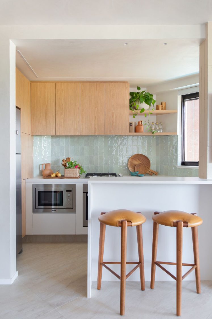 Cozinha bem iluminada com armários de madeira, revestimento verde-claro, bancada e banquedas de madeira
