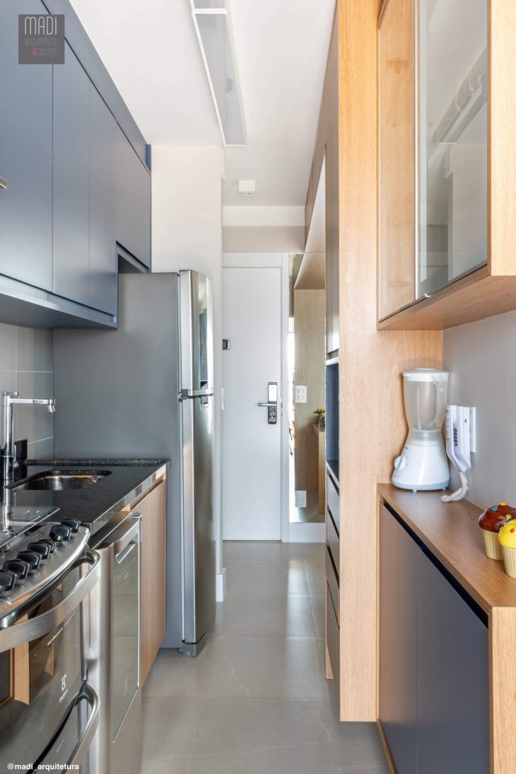 Cozinha estreita com eletrodomésticos em inox e armários com acabaento em madeira natural e azul-escuro