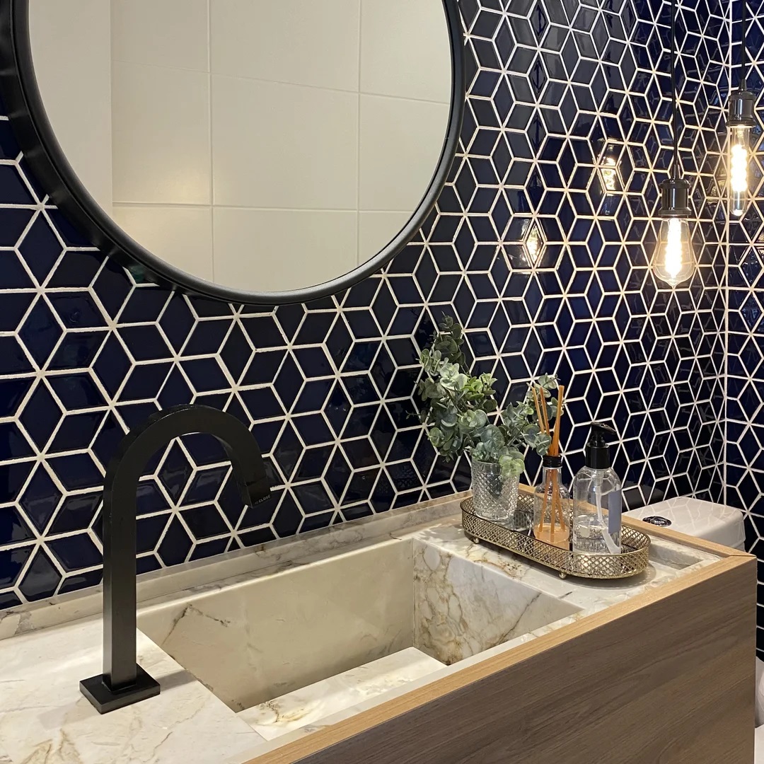 Banheiro com azulejos geométricos em azul-marinho, luminária, espelho redondo e uma bandeja com objetos na bancada