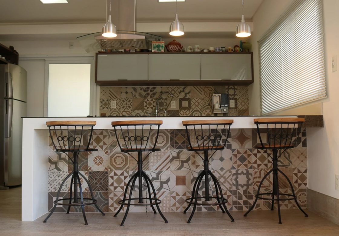Cozinha com armários claros, com acabamento em madeira, ilha revestida com ladrilhos estampados, banquetas estilo retrô e luminárias
