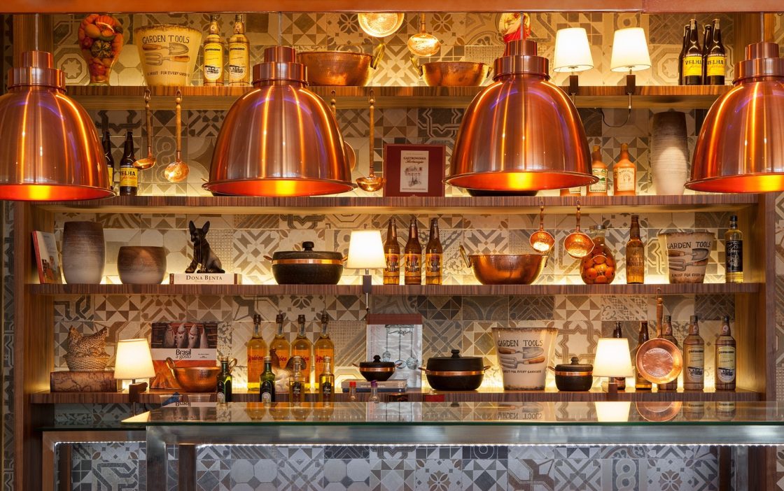 Área de restaurante retrô com azulejos estampados, luminárias acobreadas e estantes com diversos objetos e utensílios