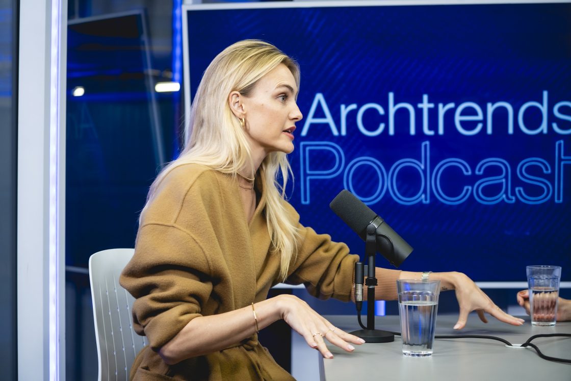 Carol Trentini em um estúdio gravando podcast, na imagem ela fala em frente a um microfone utilizando casado trench coach caramelo em um cenário azul onde se lê Archtrends Podcast