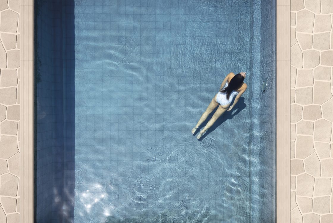 Vista aérea de piscina. Uma mulher está nadando. O fundo da piscina é azulado e as bordas são em revestimento acinzentado
