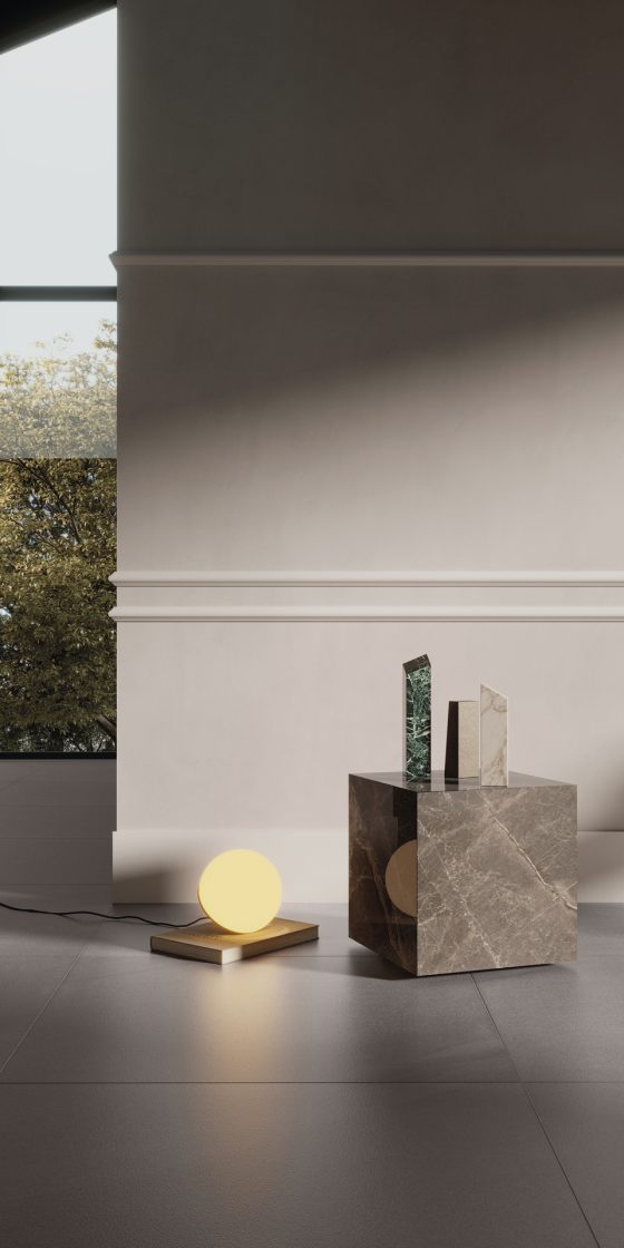 Detalhe de ambiente com parede acinzentada com rodameio, mesa de apoio com objetos e uma luminária