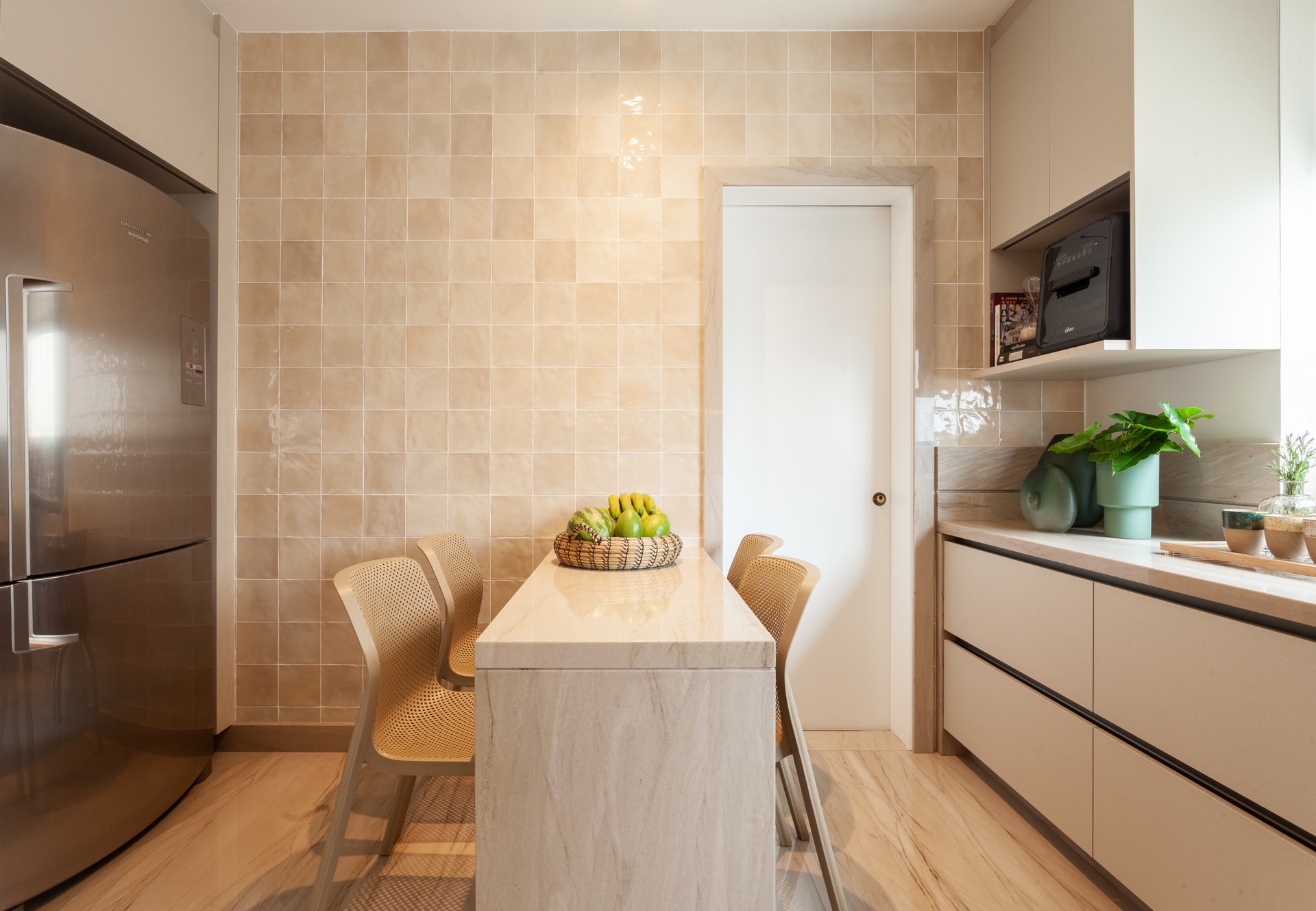 Cozinha com parede em revestimento na cor pêssego, armários claros e mesa com cadeiras. Há uma geladeira em aço inox