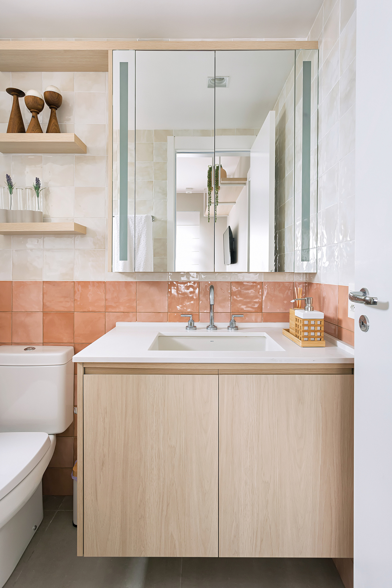 Banheiro com meia parede em revestimento na cor pêssego, armários em madeira com bancada clara, prateleiras com enfeites e espelho