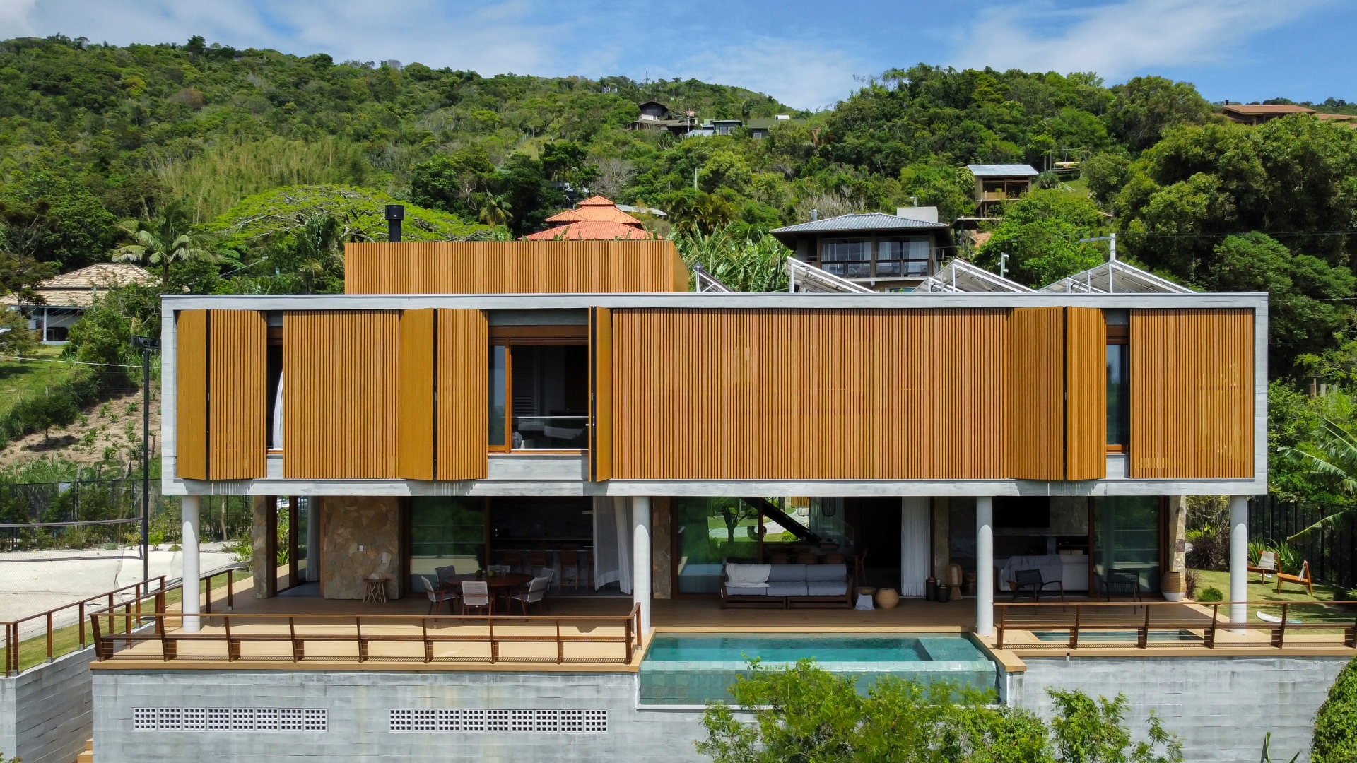 Casa de praia com fachada em madeira, dois pavimentos, piscina e área de varanda no pilotis, no telhado placas de energia solar e vegetação densa ao fundo