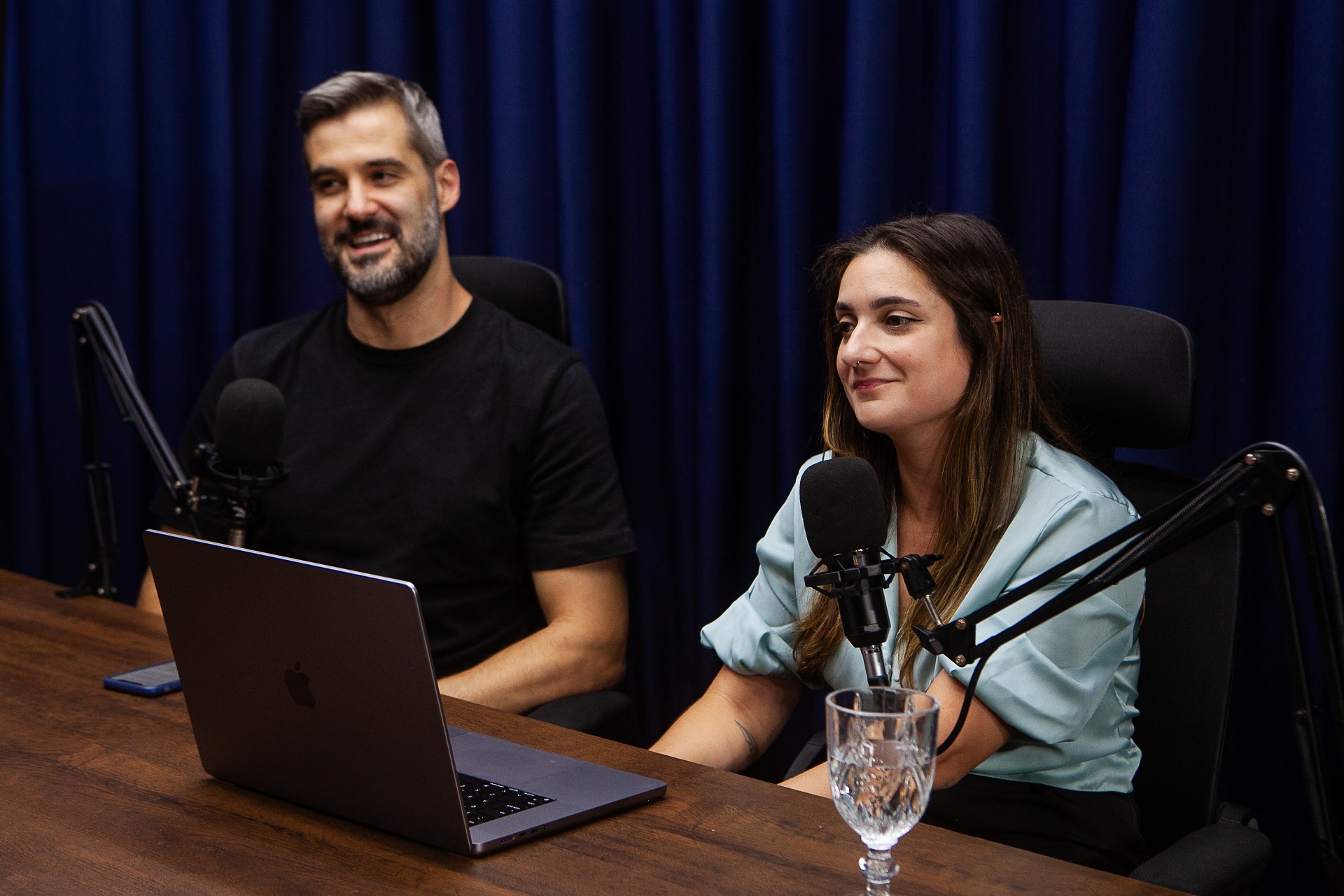 Um homem e uma mulher sentados em mesa de podcast, com microfone e notebook
