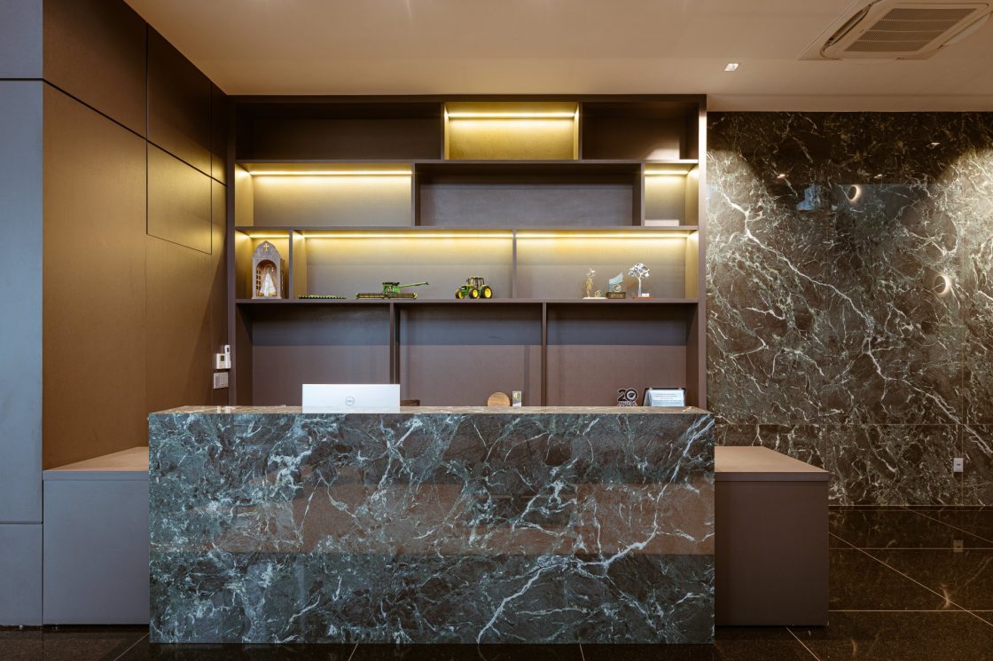Recepção com balcão em porcelanato que reproduz mármore verde, parede revestida no mesmo material e marcenaria em madeira com iluminação embutida