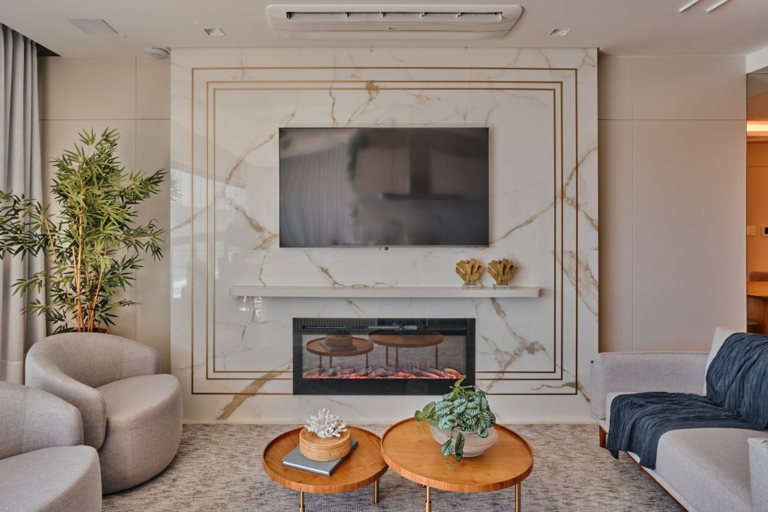 Sala de estar com televisão sobre painel de porcelanato que reproduz mármore branco e lareira abaixo