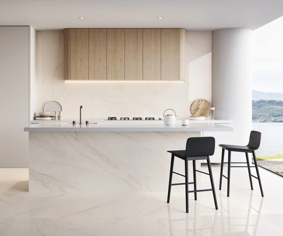Cozinha com ilha e duas banquetas pretas. Revestimentos são brancos e reproduzem Carrara