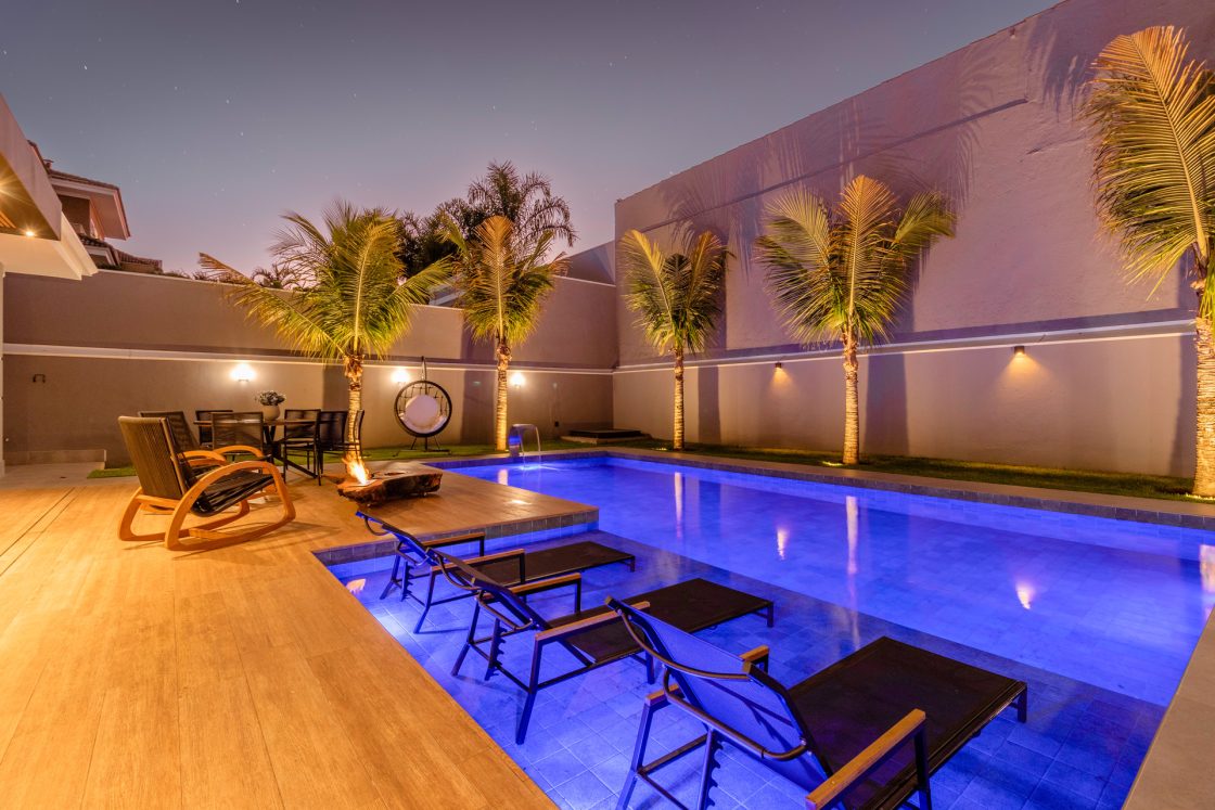 Foto noturna de área externa com piscina, espreguiçadeiras, palmeiras e spots ligados nas paredes