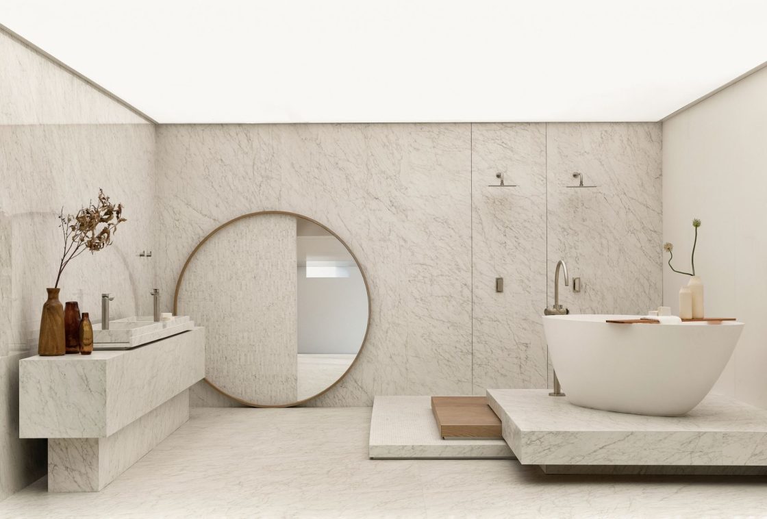 Banheiro amplo revestido em porcelanato branco com veios cinzas sutis. Inclui bancada esculpida, banheira de imersão branca e espelho redondo grande