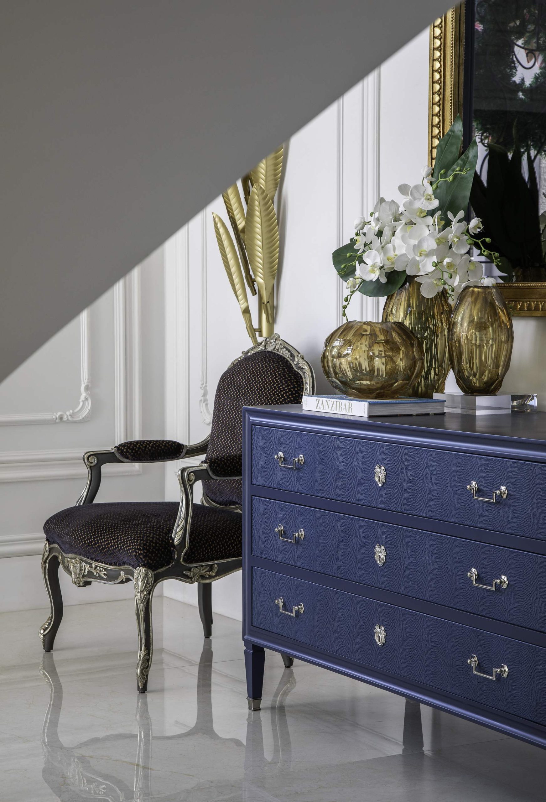 Cadeira, cômoda azul e objetos de decoração dourado compõe espaço para leitura.