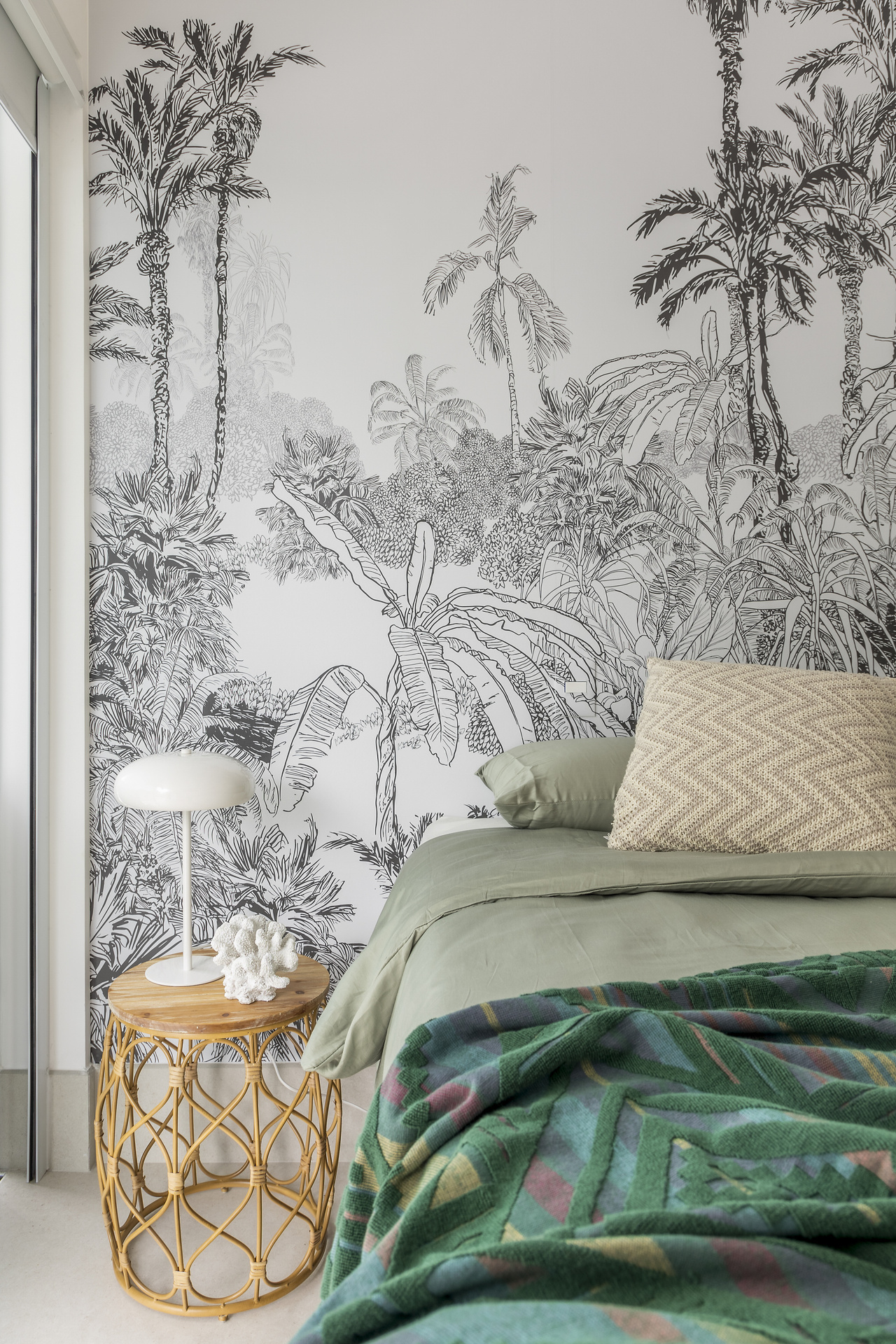 Quarto com papel de parede em preto e branco, com estampa tropical, mesa de cabeceira em madeira com abajur e cama com edredom verde.