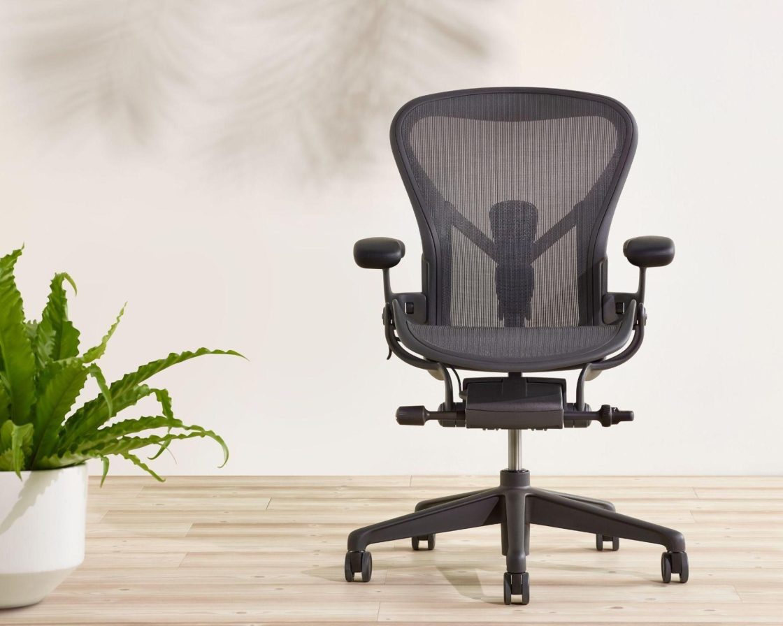 Fotografia de uma cadeira ergonômica Aeron preta ao lado de um vaso de plantas branco.