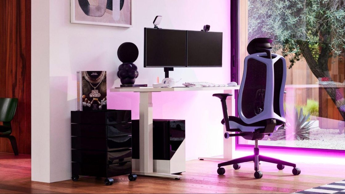 Fotografia de um espaço interno com um setup gamer que inclui uma cadeira ergonômica Vantum na cor preta e lilás, uma mesa ajustável branca e um computador gamer com dois monitores, além de itens de escritório.