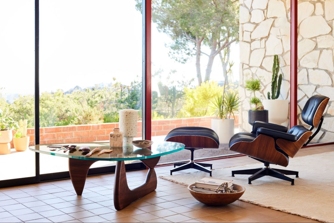 Fotografia de uma sala de estar com uma mesa, uma poltrona ergonômica com apoio para os pés, itens de decoração e uma vista para o campo através de uma porta de vidro.