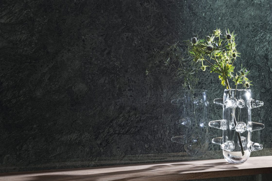 Detalhe de parede com porcelanato que reproduz mármore verde-escuro, com vaso de planta transparente em frente