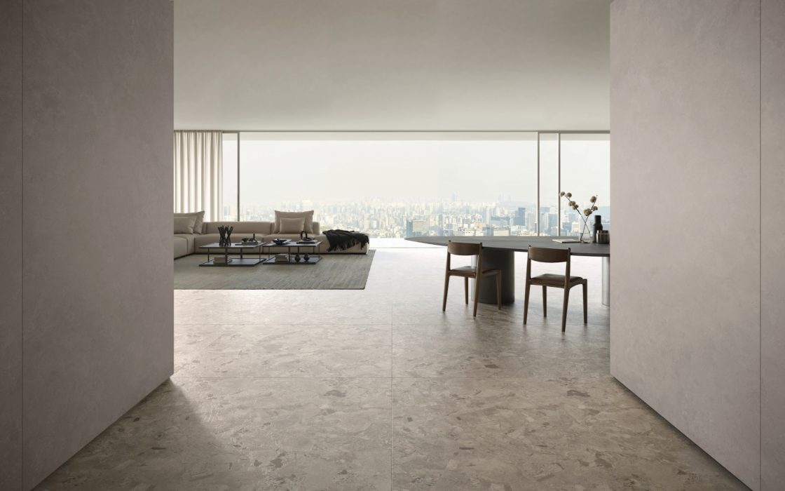 Sala de estar de apartamento, com destaque para o piso em porcelanato que reproduz breccia. O ambiente ainda conta com sofá, mesinha de centro, mesa de jantar e cadeira. Ao fundo, as grandes portas de vidro que dão para a varanda permitem ter uma visão do horizonte da cidade