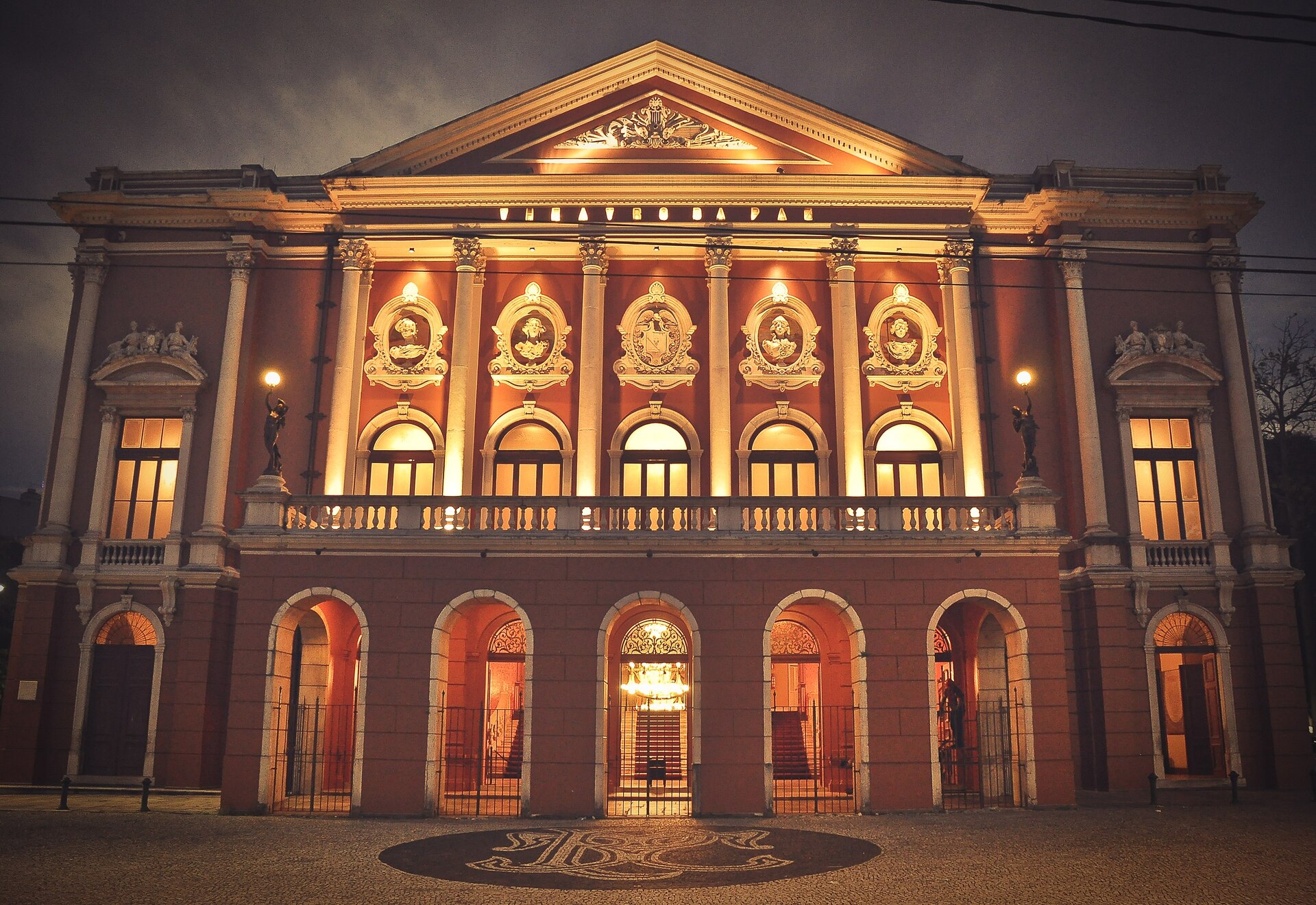 Theatro da Paz é um teatro de estilo neoclássico com dois andares, em tons de branco e terracota, com grandes pilares e portais arredondados. Foto feita à noite, com os portais iluminados